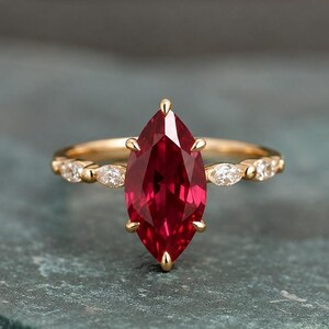 Puede incluir: Un anillo de oro con una gema roja intensa de talla marquesa como pieza central. La banda está adornada con pequeñas piedras claras. El anillo se exhibe sobre una superficie verde moteada, resaltando el diseño de la joya.
