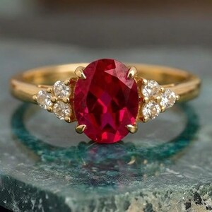 May include: A gold ring featuring a large, oval-cut ruby gemstone as the centerpiece. The ring is flanked by smaller, round, clear gemstones on each side. The ring is set on a textured, green surface, creating a striking contrast.