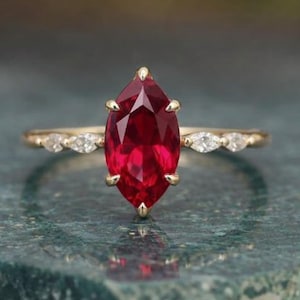 May include: A gold ring featuring a marquise-cut, vibrant red gemstone as the centerpiece. The band is adorned with small, clear accent stones. The ring is set against a blurred background, highlighting the jewelry's details.