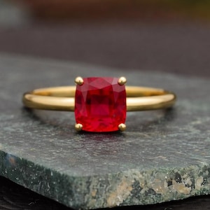 Puede incluir: Un anillo de oro con una gran gema roja brillante con talla cojín. El anillo tiene una banda de oro pulido simple, y la gema está asegurada por cuatro pequeñas garras de oro. El anillo se muestra sobre un fondo oscuro y texturizado.