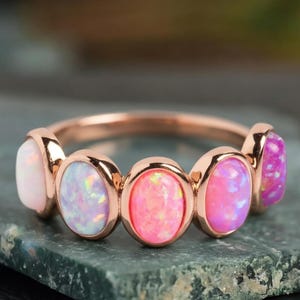 May include: A rose gold ring featuring five oval-shaped opals in shades of pink and white. The opals are set in individual bezels, creating a delicate and eye-catching design. The ring is a piece of jewelry.