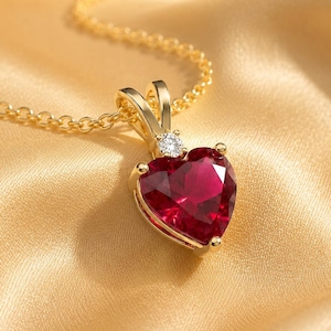 May include: A gold necklace featuring a heart-shaped red gemstone pendant. The pendant is suspended from a delicate gold chain and includes a small, clear, round-cut stone set at the top of the heart, connecting it to the chain bail.