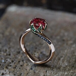 Può includere: Un anello in oro rosa con una grande gemma rossa rotonda. La gemma è circondata da pietre di accento verdi e nere. L'anello ha un design unico e fluido.