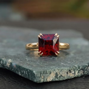 May include: A gold ring featuring a large, rectangular, deep red gemstone. The gemstone is set with four claw prongs. The ring band is a simple, polished gold. The ring is displayed on a textured, green-gray surface.