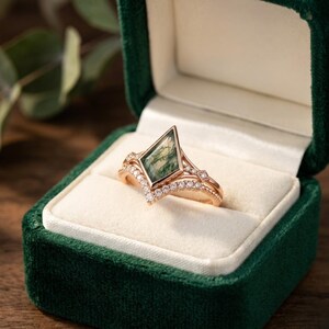 以下が含まれることがあります： 緑色のベルベットのリングボックスに入ったローズゴールドのリング。婚約指輪は、幾何学的な形のモスアゲートストーンが特徴です。バンドは、小さく輝くダイヤモンドでアクセントがつけられています。結婚指輪は、より多くのダイヤモンドが付いた曲線的なデザインです。