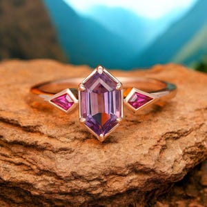 May include: A rose gold ring featuring a large octagon-shaped amethyst gemstone, flanked by two smaller triangular ruby gemstones.