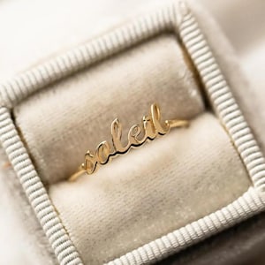 May include: A gold-colored ring with the word "soleil" in cursive script. The ring is displayed inside a gray velvet jewelry box. The ring's delicate design and warm color create an elegant aesthetic.