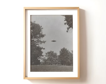 May include: A black and white photograph of a flying saucer hovering over a field of trees. The photo is framed in a light wood frame.