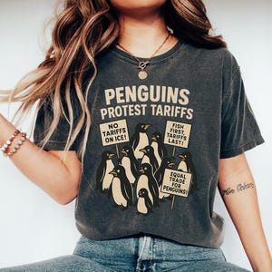 Könnte beinhalten: Ein dunkelgraues T-Shirt mit einer Grafik von Pinguinen, die Schilder halten, auf denen steht: "Keine Zölle auf Eis!" und "Fisch zuerst, Zölle zuletzt!" und "Gleicher Handel für Pinguine!"
