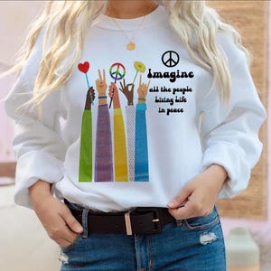 Op de afbeelding: Witte sweatshirt met de tekst "Imagine all the people living life in peace". De afbeelding toont kleurrijke armen die vredessymbolen, een hart en een bloem vasthouden. De sweatshirt heeft lange mouwen en een ronde hals.
