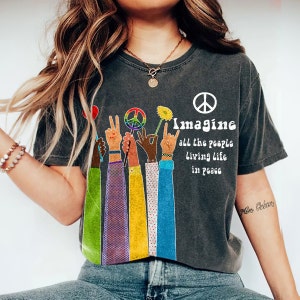 Op de afbeelding: Een donkergrijs T-shirt met een vredesteken en de tekst "Imagine all the people living life in peace". Het ontwerp bevat kleurrijke handen die vredestekens, een bloem en een hart vasthouden.