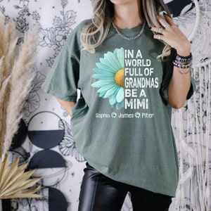 May include: Green t-shirt with a blue daisy graphic and the text "In a world full of grandmas be a mimi". The shirt also has the names "Sophia", "James", and "Piter" printed below the graphic.