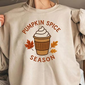 May include: Beige sweatshirt with an embroidered design featuring a coffee cup with whipped cream, autumn leaves, and the text "PUMPKIN SPICE SEASON". The word "EMBROIDERED" is at the bottom.