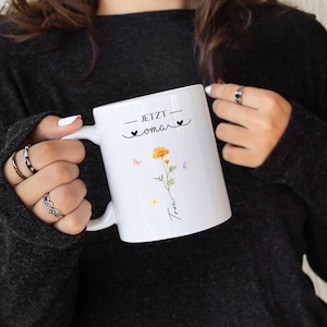 May include: White ceramic mug with the text "- JETZT omar -" and a floral design featuring a yellow flower and butterflies. The mug has a curved handle and is held by a person wearing a black sweater.