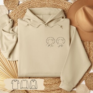 Könnte beinhalten: Beigefarbener Kapuzenpullover mit Kordelzug und Vordertasche. Der Pullover hat ein minimalistisches Strichdesign von zwei Gesichtern mit den Worten "Mimi" und "Papa" darunter. Ein Hut und ein gewebter Hintergrund sind ebenfalls zu sehen.