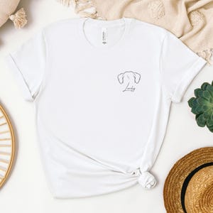 May include: White t-shirt featuring a minimalist black outline of dog ears and the word "Lucky" in script. The shirt has short sleeves and a crew neck. The shirt is made of a soft material.