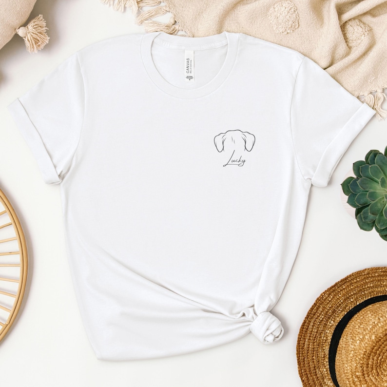 May include: White t-shirt featuring a minimalist black outline of dog ears and the word "Lucky" in script. The shirt has short sleeves and a crew neck. The shirt is made of a soft material.