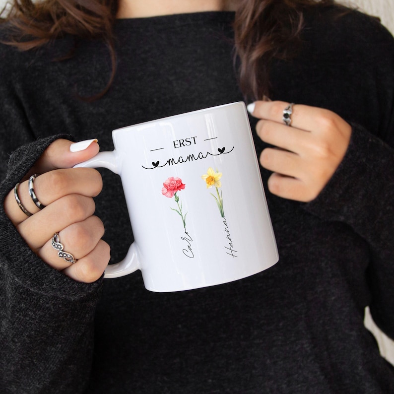 May include: White ceramic mug with the text "ERST mama" and two floral illustrations. One illustration is a red carnation, and the other is a yellow daffodil. The names "Caro" and "Hanna" are written below the flowers.