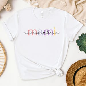 Könnte beinhalten: Weißes T-Shirt mit dem Wort "mami" in farbenfrohen, stilisierten Buchstaben. Das Wort ist in Rosa, Lila, Gelb und Orange gehalten, mit kleinen Herz- und Punktakzenten. Das Shirt besteht aus einem weichen Material.