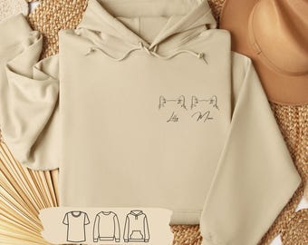 Könnte beinhalten: Ein beiger Hoodie mit einer minimalistischen Linienzeichnung von zwei Katzenköpfen und den Namen "Lilly" und "Mimi" darunter. Der Hoodie ist auf einem geflochtenen Strohhintergrund mit einem braunen Hut und einer Auswahl anderer Kleidungsstücke zu sehen: ein T-Shirt, ein Sweatshirt mit Rundhalsausschnitt und ein Hoodie.