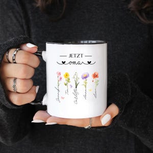 May include: White ceramic mug with a black interior and handle. The mug features the text "JETZT oma" with floral illustrations and names. The design includes colorful flowers and names in a script font, perfect for a personalized gift.