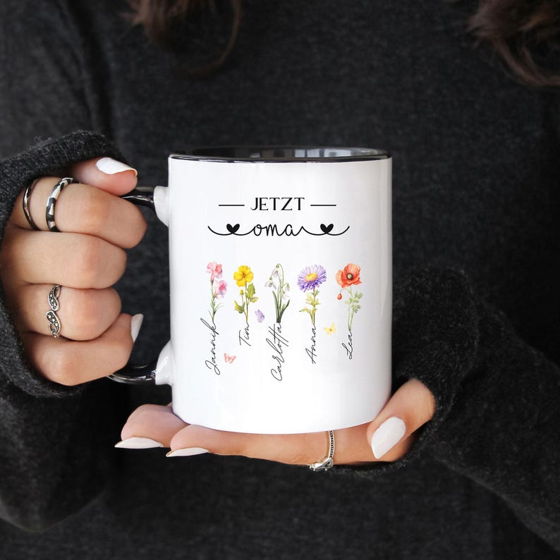 May include: White ceramic mug with a black interior and handle. The mug features the text "JETZT oma" with floral illustrations and names. The design includes colorful flowers and names in a script font, perfect for a personalized gift.