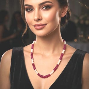 May include: A beaded necklace featuring alternating round, deep magenta-colored beads and smaller, smooth white spherical beads. The necklace is worn around the neck, showcasing a repeating pattern of the dark pink and white beads in a single strand.