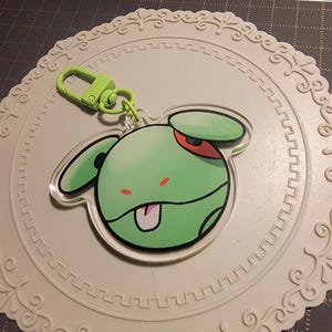May include: A green cartoon character keychain with a green clasp. The character has a green head, ears, and a red eye. The keychain is on a white, decorative, circular coaster.
