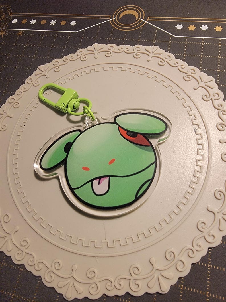 May include: A green cartoon character keychain with a green clasp. The character has a green head, ears, and a red eye. The keychain is on a white, decorative, circular coaster.