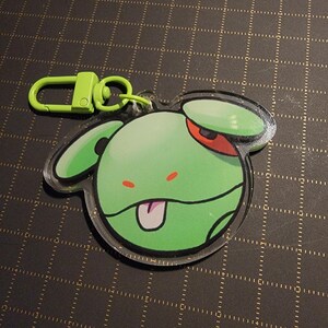 May include: A green cartoon character keychain with a lime green clasp. The character has a green body, red eye, and a pink tongue sticking out. The keychain is made of clear acrylic.