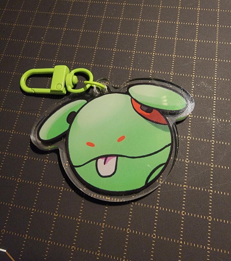 May include: A green cartoon character keychain with a lime green clasp. The character has a green body, red eye, and a pink tongue sticking out. The keychain is made of clear acrylic.