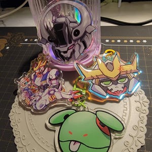 May include: Collection of anime-style acrylic keychains featuring robot and character designs. The keychains have vibrant colors and holographic accents. One keychain has a green character with its tongue out.