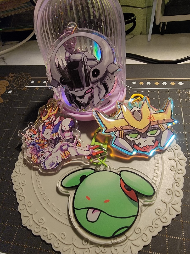 May include: Collection of anime-style acrylic keychains featuring robot and character designs. The keychains have vibrant colors and holographic accents. One keychain has a green character with its tongue out.