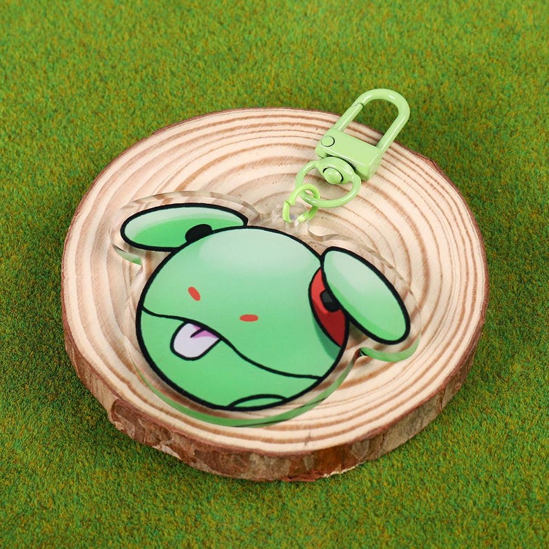 May include: A green cartoon character keychain with a green clasp. The character has a green body, red eye, and a white tongue sticking out. The keychain is on a wooden slice.