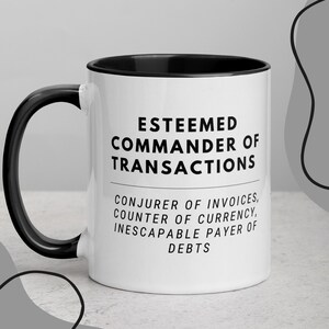 以下が含まれることがあります： 黒色の縁取りと取っ手を持つ白いセラミック製のコーヒーカップ。カップには、黒い文字で「Esteemed Commander of Transactions, Conjurer of Invoices, Counter of Currency, Inescapable Payer of Debts.」と書かれています。