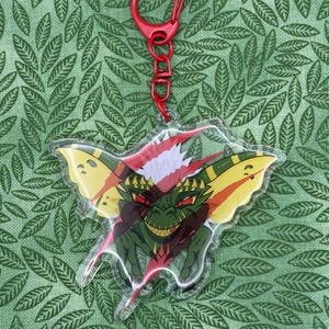May include: A clear acrylic keychain featuring a green creature with large yellow ears, red eyes, and sharp teeth, attached to a red plastic lobster clasp. The character has a spiky white tuft on its head and is depicted with a mischievous grin.