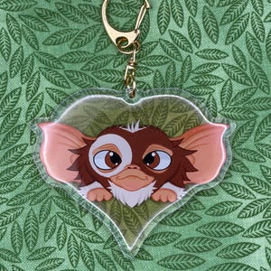 May include: A heart-shaped acrylic keychain featuring a colorful illustration of a brown and white creature with large ears and dark eyes. The keychain includes a gold-toned metal ring and clasp attached to the top of the heart-shaped acrylic piece.
