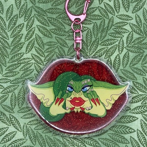 May include: A clear acrylic keychain featuring a glittery red lip-shaped background with a green-skinned creature character. The character has long green hair, blue eyeshadow, red lipstick, and sharp red fingernails. It includes a pink metal clasp.