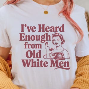 Puede incluir: Camiseta blanca con un gráfico rojo que dice "He oído suficiente de los viejos blancos". El gráfico presenta a una mujer al teléfono con un dedo en los labios.
