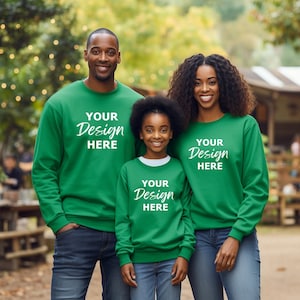 May include: A family of three wearing matching green sweatshirts with the text "YOUR DESIGN HERE" printed on them. The father is wearing blue jeans, the mother is wearing blue jeans, and the child is wearing blue jeans.