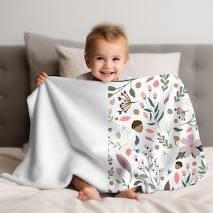 May include: A white baby blanket with a floral pattern on one side. The pattern features pink, green, and brown flowers and leaves on a white background. A baby is sitting on a white bed holding the blanket.