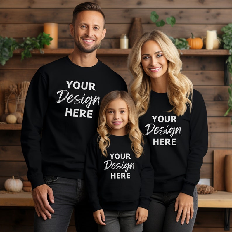 K&ouml;nnte beinhalten: Eine dreik&ouml;pfige Familie tr&auml;gt schwarze Sweatshirts mit dem Aufdruck "Your Design Here" auf der Vorderseite. Die Familie steht vor einer Holzwand.