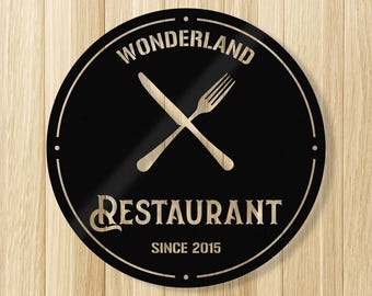 Pode incluir: Uma placa redonda de metal preto com as palavras "WONDERLAND RESTAURANT" em branco, com um design de faca e garfo cruzados. A placa também inclui o texto "SINCE 2015". Fundo de madeira.