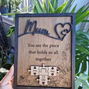 May include: Wooden plaque with the word "Mum" and a heart design. The plaque includes the text "You are the piece that holds us all together" and puzzle pieces with names. The frame is dark brown, and the background is a light wood grain.