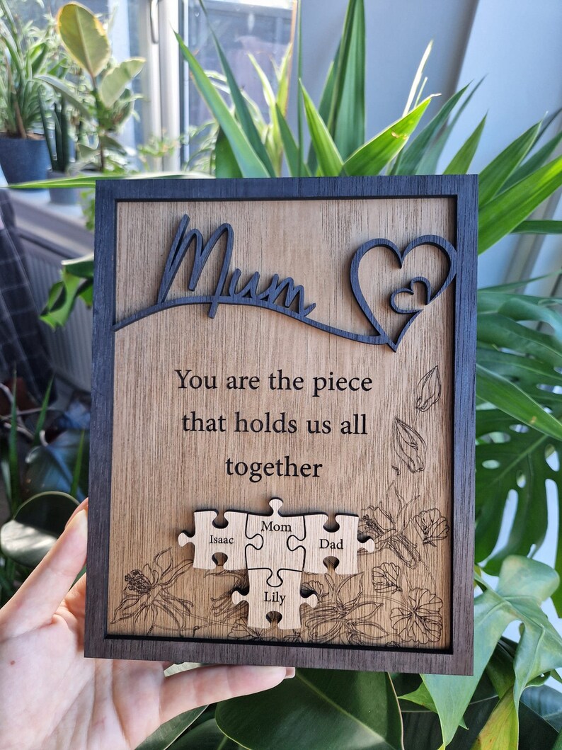 May include: Wooden plaque with the word "Mum" and a heart design. The plaque includes the text "You are the piece that holds us all together" and puzzle pieces with names. The frame is dark brown, and the background is a light wood grain.