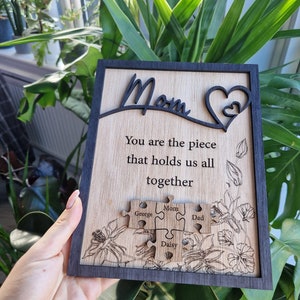 May include: Wooden plaque with a black frame and a light wood grain center. The plaque features the word "Mom" in a cursive font, a heart design, and the quote "You are the piece that holds us all together." Below are puzzle pieces with names.