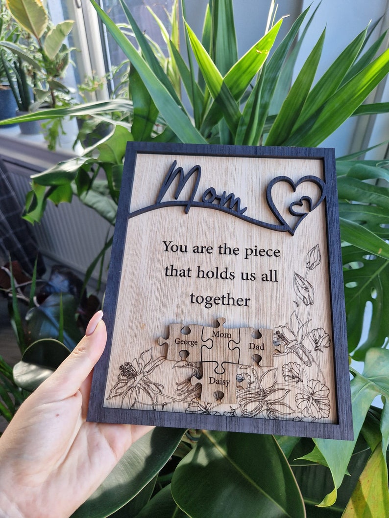 May include: Wooden plaque with a black frame and a light wood grain center. The plaque features the word "Mom" in a cursive font, a heart design, and the quote "You are the piece that holds us all together." Below are puzzle pieces with names.