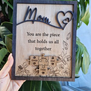 May include: Wooden plaque with a dark brown frame and a light wood interior. The word "Mom" is cut out at the top, with a heart design. Text reads "You are the piece that holds us all together." Puzzle pieces are engraved with names.