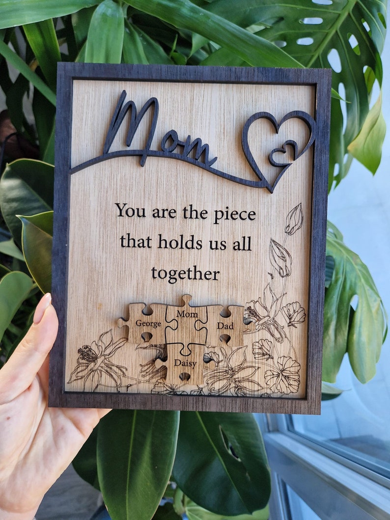 May include: Wooden plaque with a dark brown frame and a light wood interior. The word "Mom" is cut out at the top, with a heart design. Text reads "You are the piece that holds us all together." Puzzle pieces are engraved with names.