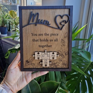 May include: Wooden plaque with a dark frame and a light wood grain center. The word "Mum" is cut out at the top, with a heart design. The text reads "You are the piece that holds us all together" with puzzle pieces below.